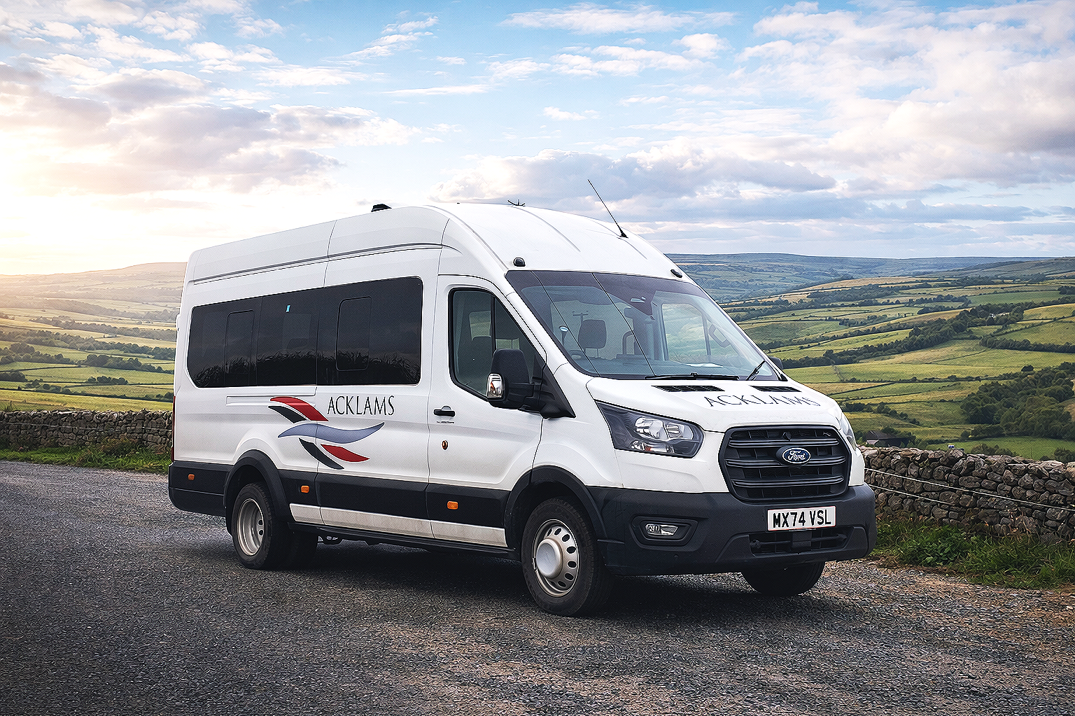 A white Ford transit-style van with "ACKLAM" written on the side, parked on a rural road with green rolling hills and cloudy sky in the background.