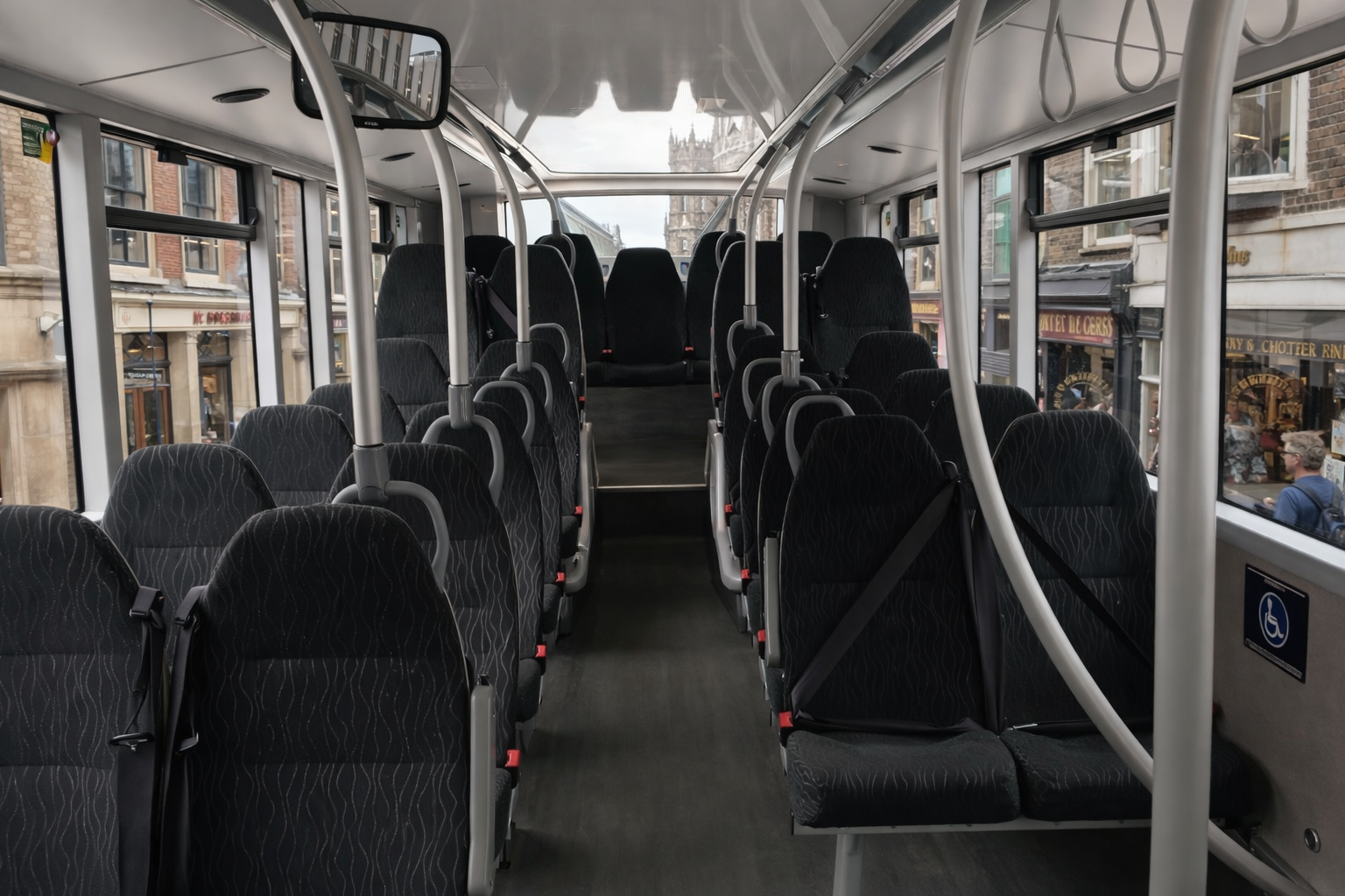 Interior of a bus with empty black fabric seats, large windows, and a view of a city street with buildings and pedestrians outside.