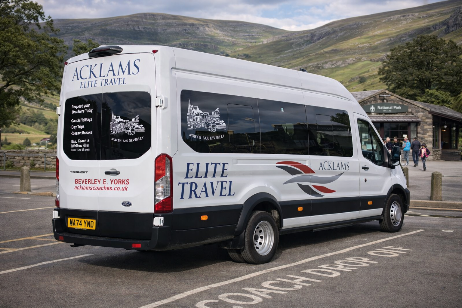 White Elite Travel minibus parked in a lot near a stone building with a sign for a national park, with mountains and trees in the background and a group of people walking nearby.