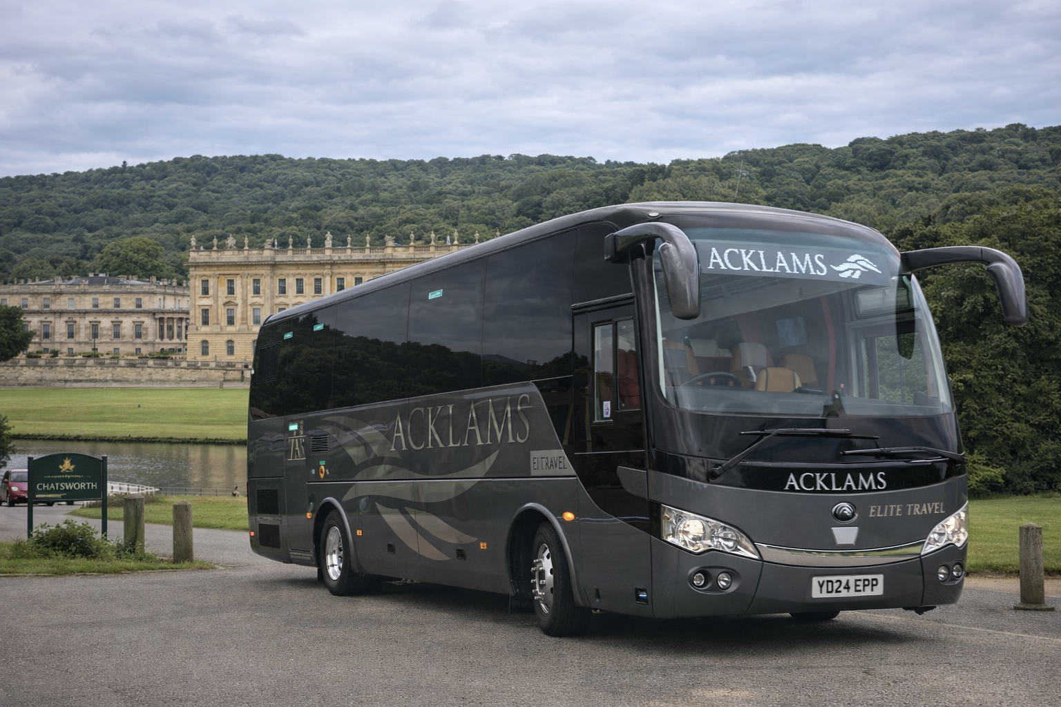 A black luxury tour bus with 'ACKLAMS' and 'ELITE TRAVEL' written on it, parked outdoors near a body of water and historic building with greenery and hills in the background.