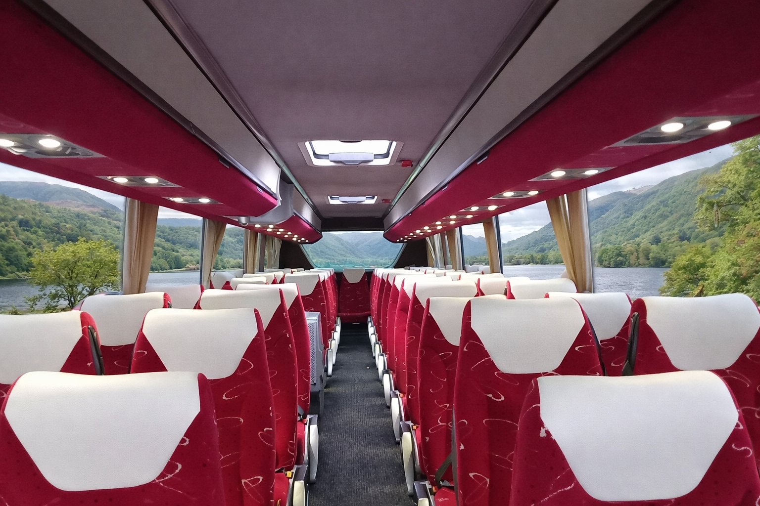 Interior of a bus with red and white seats, large windows showing a scenic view of mountains and a lake, and overhead storage compartments.