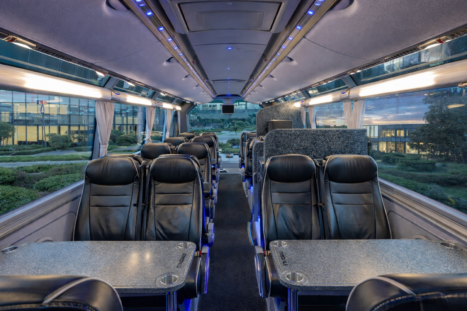 Empty luxury bus interior with black leather seats, small tables, large windows with white curtains, and blue ambient lighting.