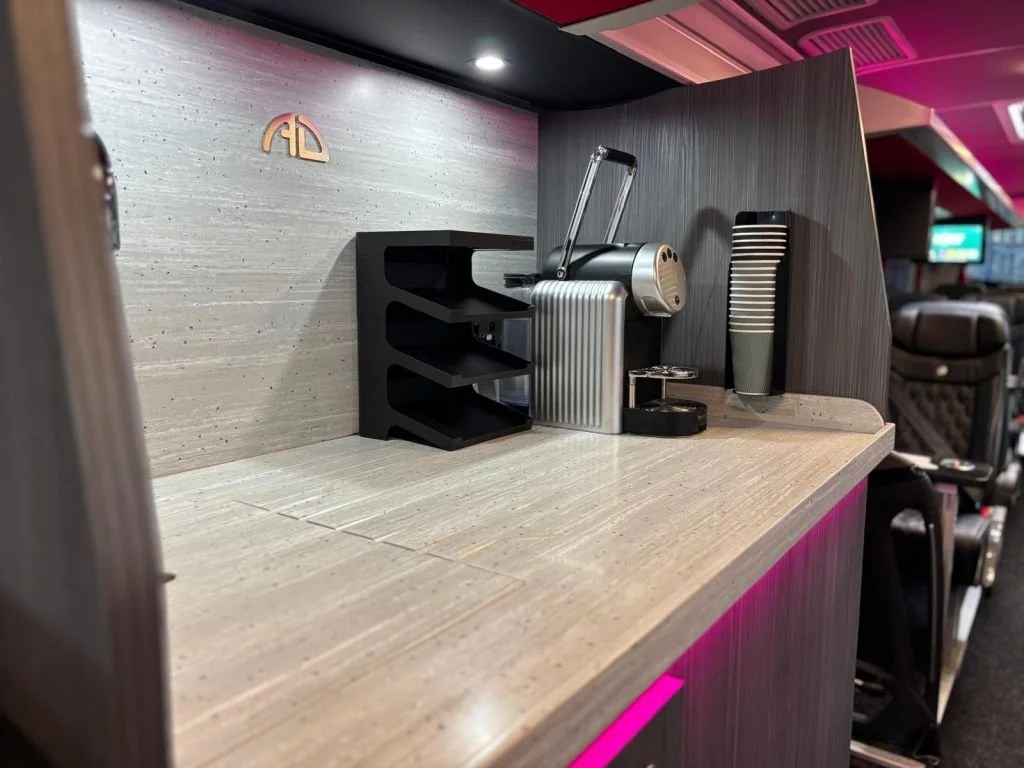 Empty airport or lounge counter with a small black shelf, a silver coffee maker, a black thermos, and a black box, with a textured wall behind and a seat in the background.