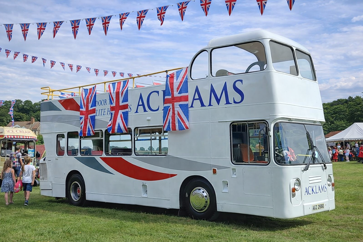 A double-decker bus decorated with British flags and the word 'ACKLAMS' on the front is parked on a grassy field during a celebration. People are gathered around, and there are bunting flags flying in the sky.