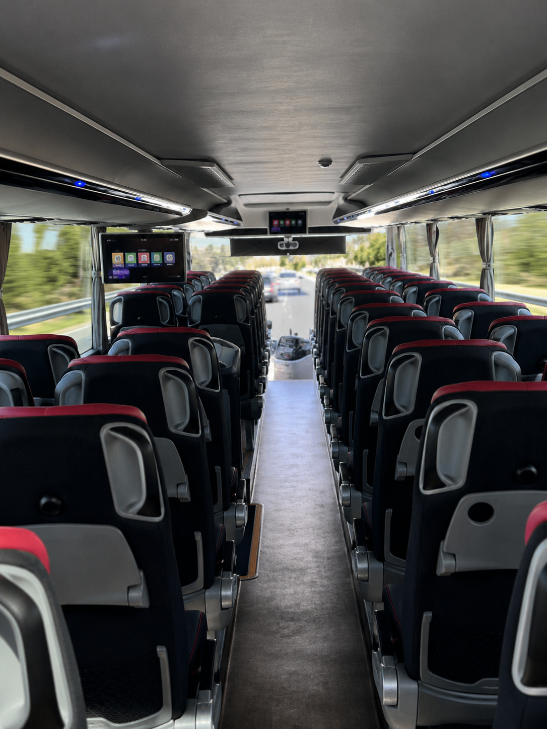 Inside view of a modern bus with black and red seats, overhead entertainment screens, and large windows showing a highway and trees outside.