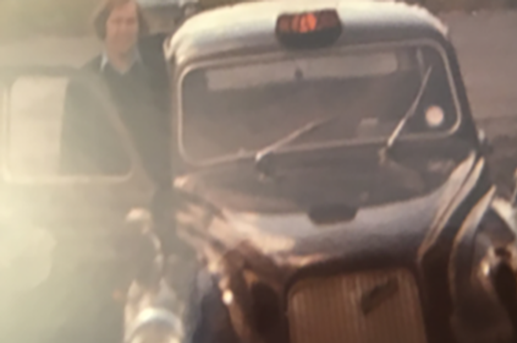 A person standing next to a vintage black car with a rounded hood and large front grille.