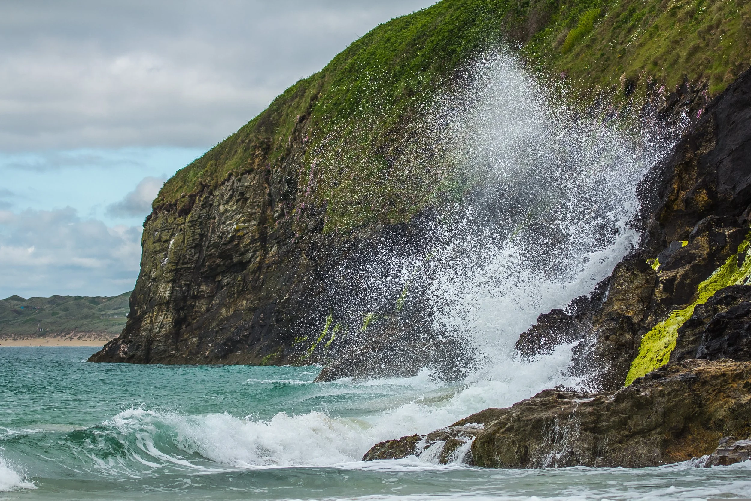 Waves crashing against a rocky cliff on a cloudy day with green vegetation on the cliffside.