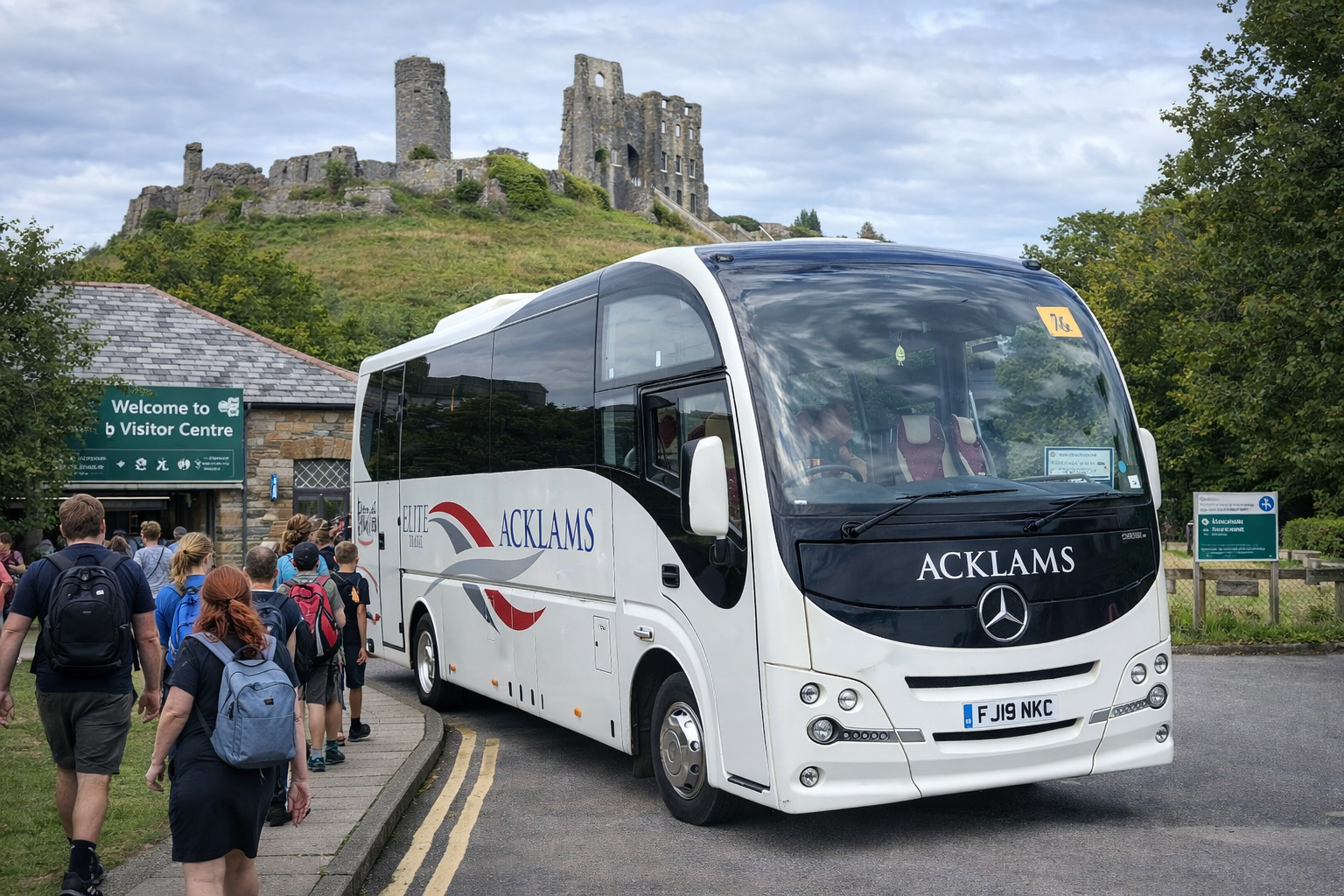 Tour bus with the word 'ACKLAMS' on the front and side, parked at a visitor center with tourists walking in line. There is a castle on a hill in the background and trees around.