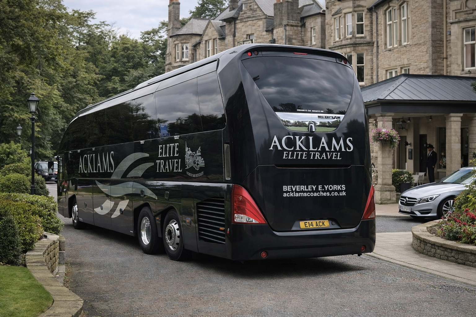 A large black coach bus with the words 'Acklams Elite Travel' parked in front of a stone building with a porch. There are trees, a lamp post, a silver car, and a man in a uniform near the entrance of the building.