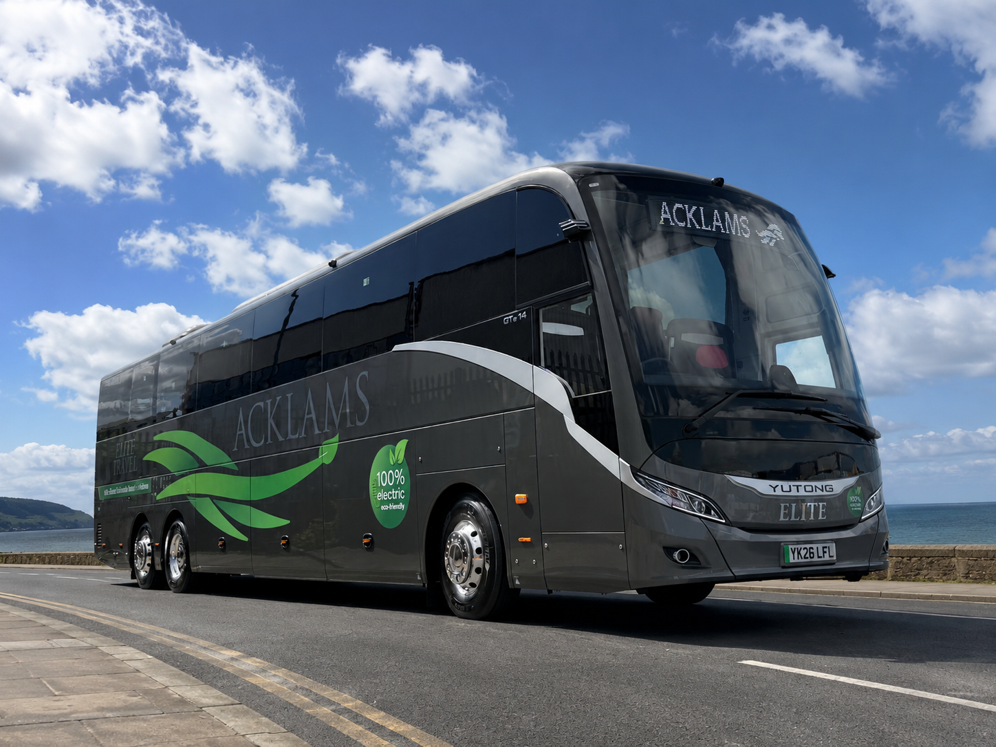 A sleek, grey electric bus with the name 'ACKLAMS' and 'YUTONG ELITE' on its front, driving along a seaside road under a blue sky with scattered clouds.