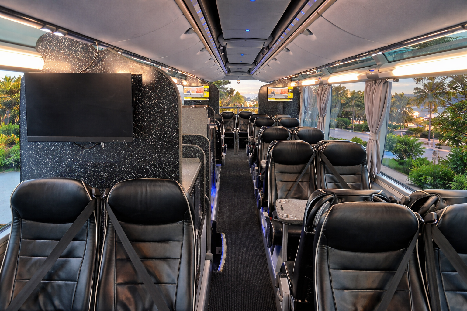 Inside a bus with black leather seats, large windows with curtains, and multiple TV screens, during either sunrise or sunset, with a view of trees, bushes, and a parking lot outside.