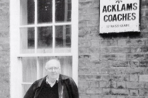 A man standing outside in front of a brick wall with a window and a sign that reads 'ACKLAMS COACHES' with a phone number.