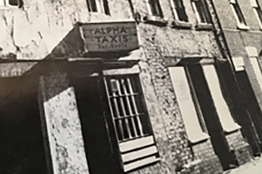 A black and white photo of a building with a sign that reads 'ALPHA TAXIS' above the door.