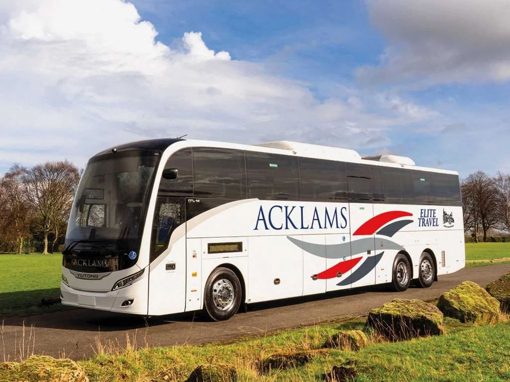 White tour bus with 'ACKLAMS' and 'Elite Travel' written on the side, parked on a paved road in a grassy area with trees and cloudy sky in the background.