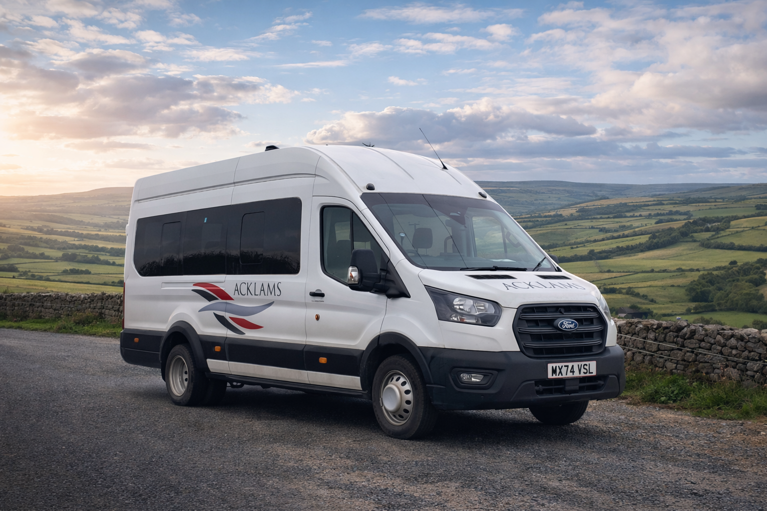 White passenger van with 'ACKLAMS' logo parked on a rural road with rolling green hills and a cloudy sky in the background.