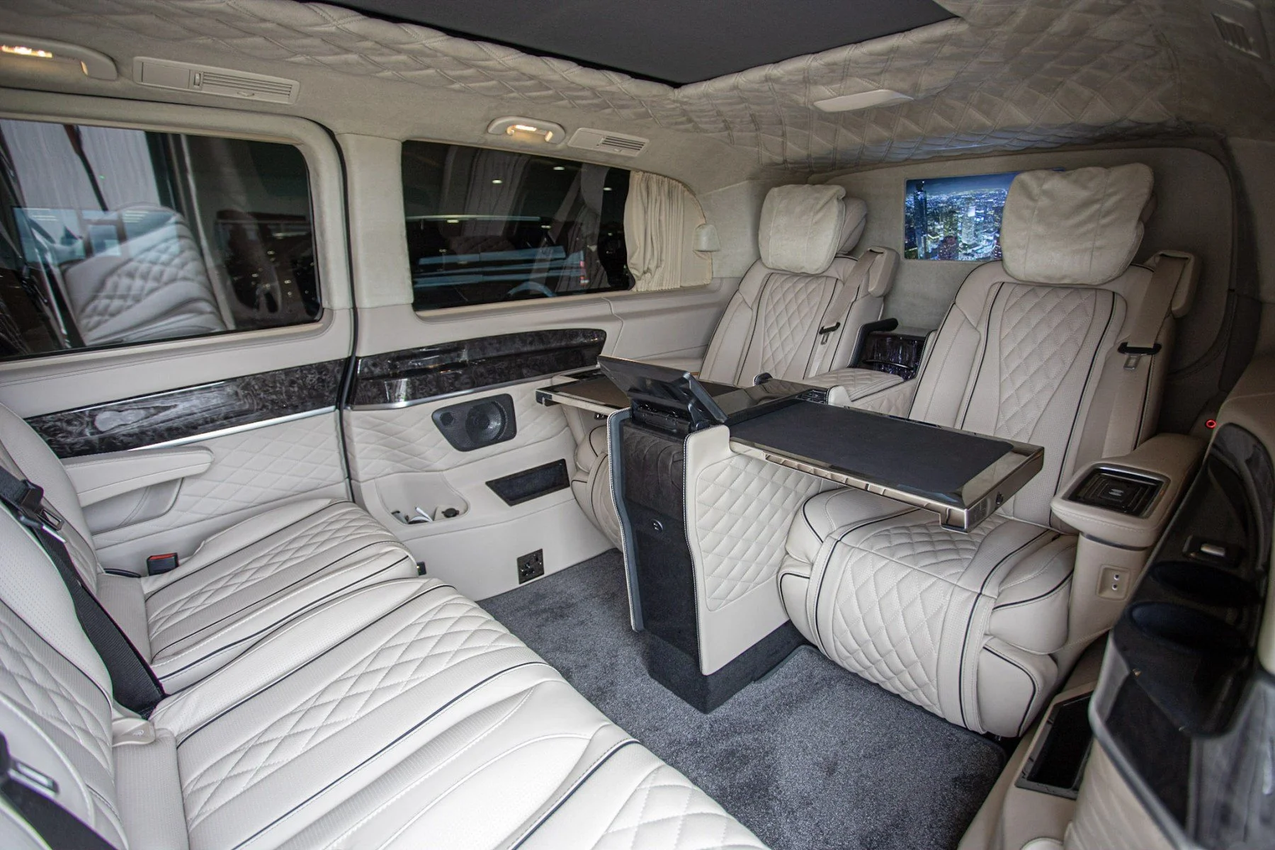 Interior of a luxurious vehicle with cream-colored quilted leather seats and built-in entertainment options, viewed from the side window showing a cityscape at night.