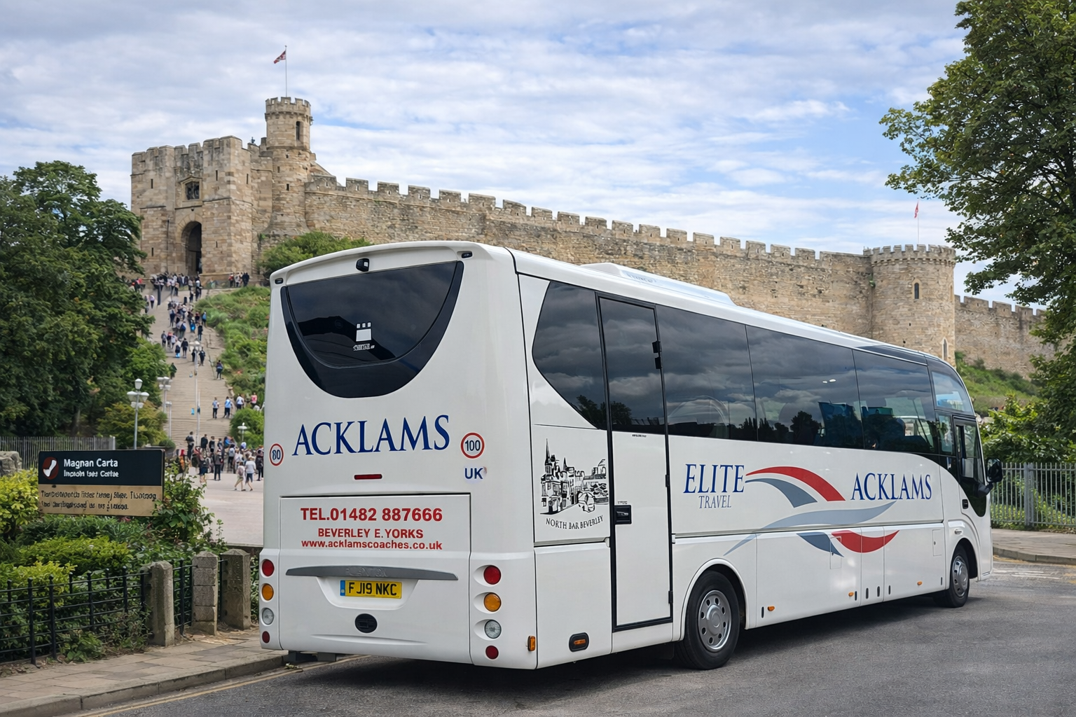 A white coach bus with blue and red logo markings parked near a castle with a stone wall and towers, alongside a sign for Magnan Carta bus services.