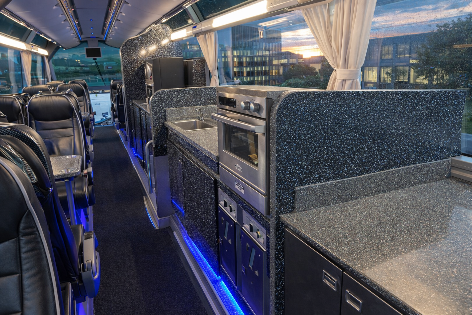 Interior of a luxury bus with black leather seats, a small kitchen area with a sink, oven, and cabinets, and large windows with white curtains showing a cityscape and sunset outside.
