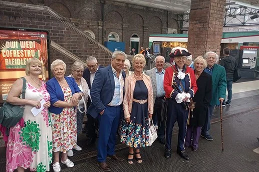 Group of elderly people, some dressed as historical figures, standing on a train platform.