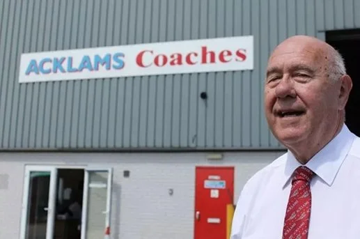 An older man in a white shirt and red tie standing outside a building with a sign that reads 'ACKLAMS Coaches'.