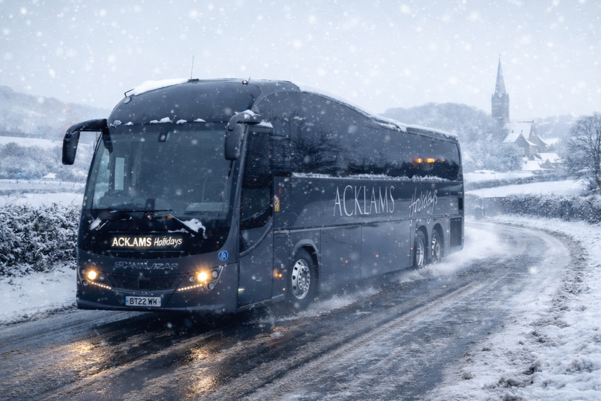A black tour bus driving through snowy, icy roads in winter with snow falling around. The bus has the words "ACKAMS Holidays" on it and headlights on.