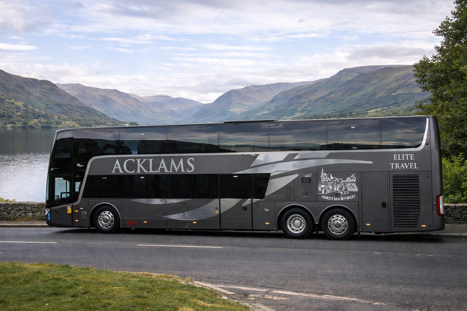 A black tour bus with 'ACKLAMS' and 'ELITE TRAVEL' written on it, parked on a road beside a lake with mountains in the background.