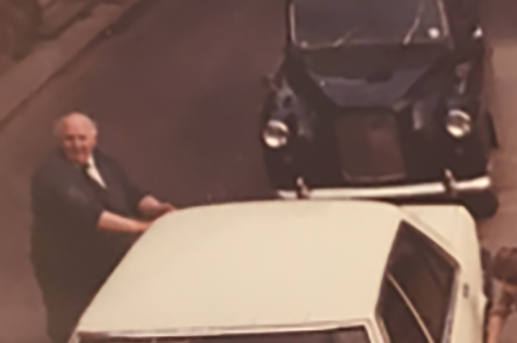 An elderly man smiling and standing beside a white car on a street, with another black car parked behind him.