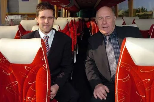 Two men sitting inside a bus with red patterned seats, smiling at the camera.