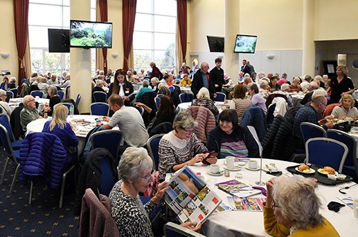 A large room filled with many elderly people seated at round tables enjoying a meal, with some reading magazines. The room is well-lit with large windows, and multiple flat-screen TVs are mounted on pillars around the room.