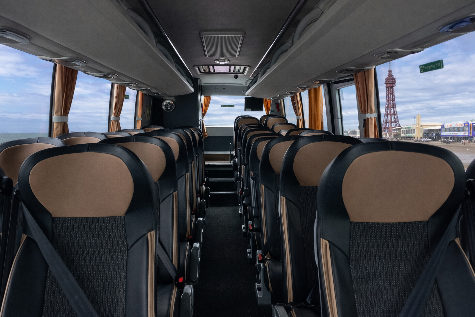 Interior of an empty bus with beige and black seats, large windows with orange curtains, and a view of the coastline and a tower outside.