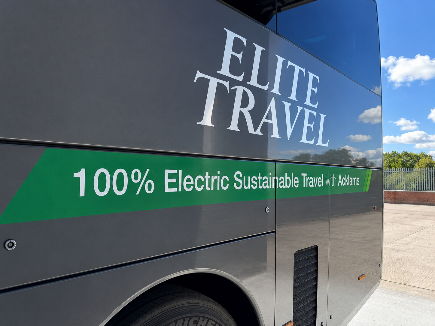 A grey Elite Travel bus with the words '100% Electric Sustainable Travel with Acklams' written on the side, parked outdoors with a bright blue sky and white clouds