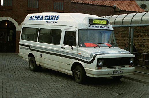 White minibus with 'ALPHA TAXIS of BEVERLEY' written on top, parked on a brick sidewalk near brick buildings, with a yellow destination sign reading 'BEVERLEY' and a black sign saying 'ACKLAM'S COACHES' in the front window.