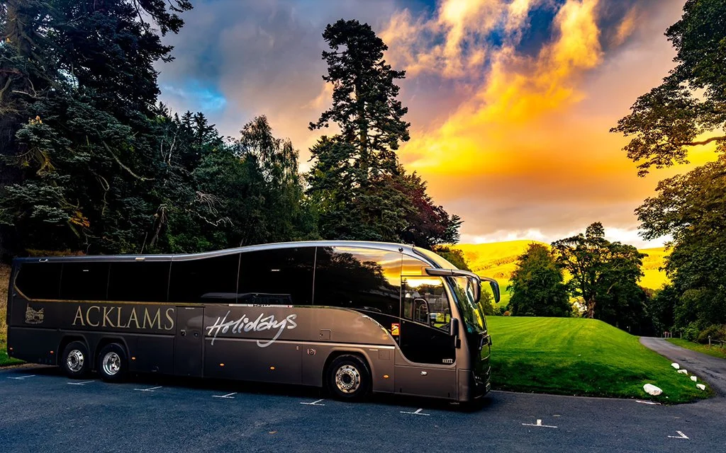 Black tour bus parked on a paved lot near green trees and grass with a scenic sunset in the background.