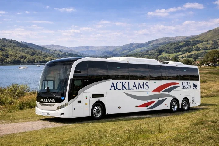Large white coach bus with the words 'ACKLAMS' and 'ELITE TRAVEL' parked on grassy terrain near a body of water, with hills and blue sky in the background.