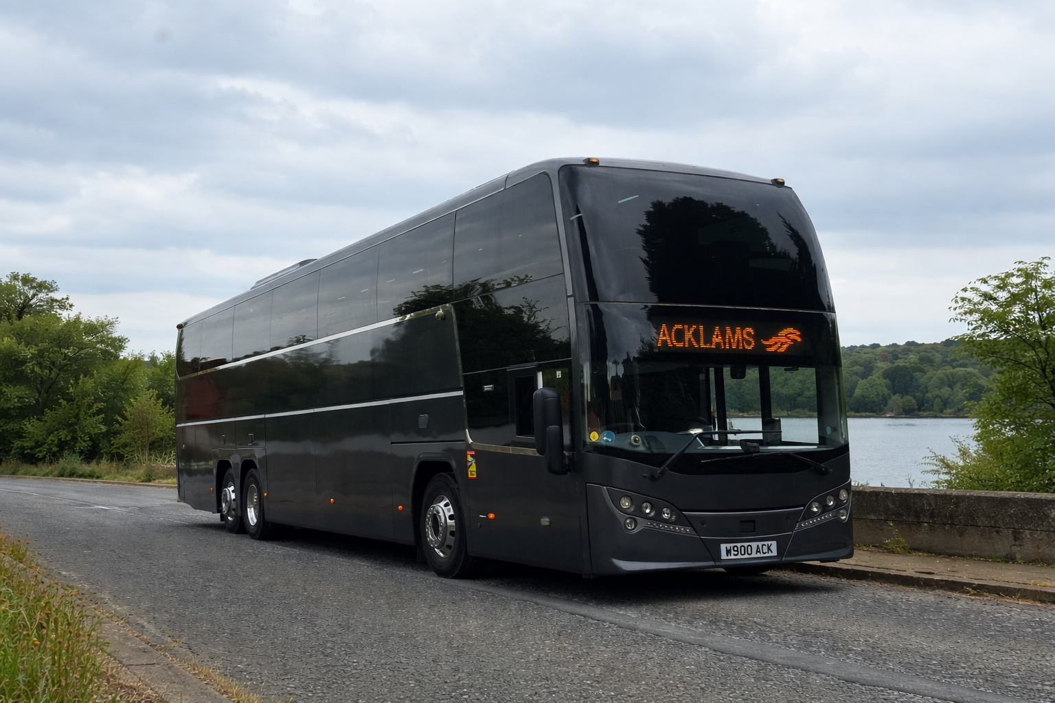 A black double-decker bus parked on the side of a road near a body of water, with greenery and hills in the background, and an electronic sign displaying the destination 'ACKLAMS'.