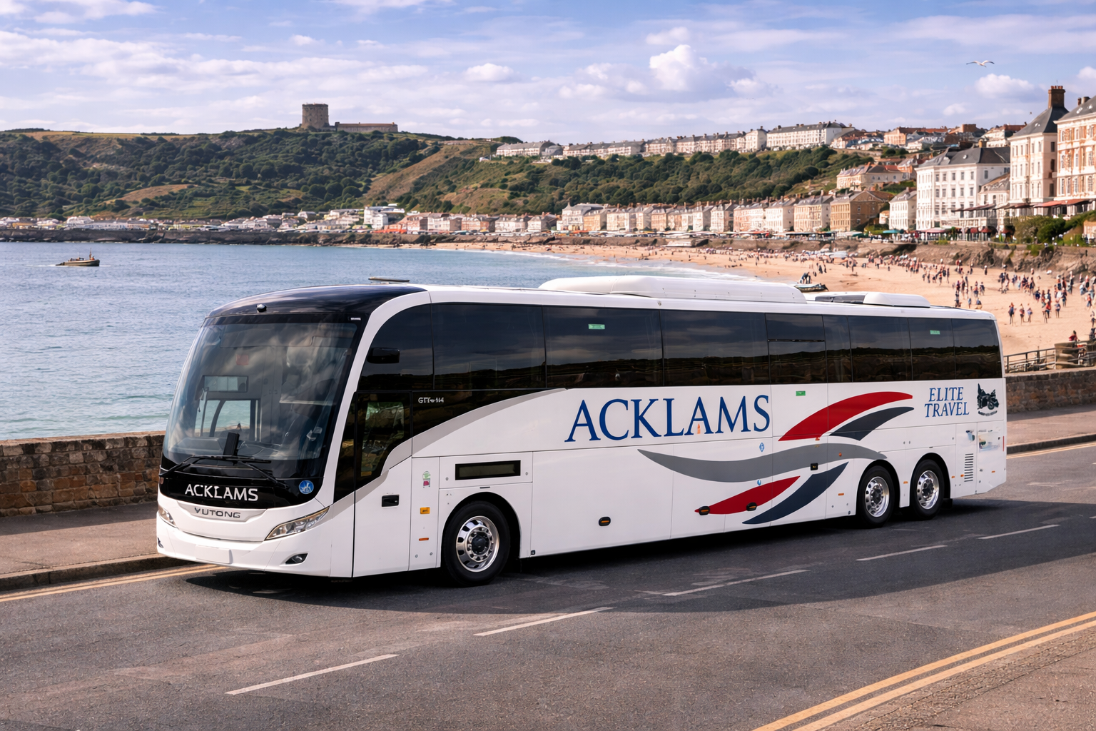 A white coach bus with 'ACKLAMS' and 'ELITE TRAVEL' logos parked on the side of a coastal road. The background features a sandy beach filled with people, a calm body of water, and a hillside with houses and a castle.