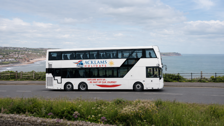 A 98 seat white double-decker bus with the logo of Acklam Holidays, traveling along a coastal road with the ocean and cliffs in the background.