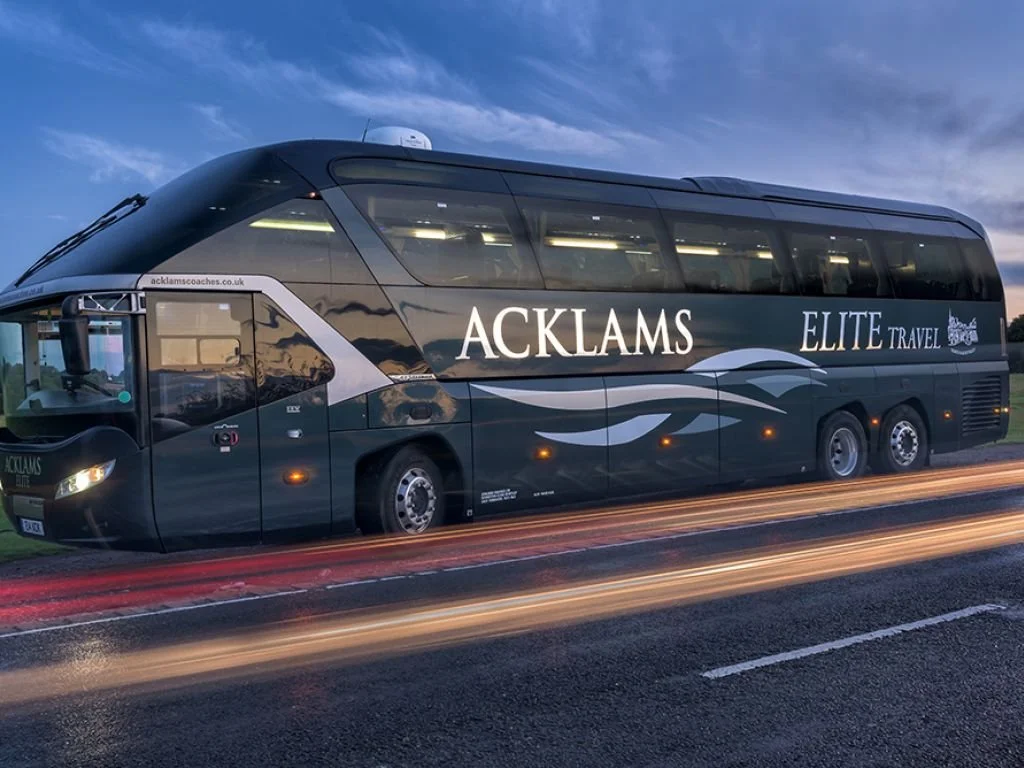 A black coach bus with the words "ACKLAMS ELITE TRAVEL" on the side, parked on the side of the road during dusk.