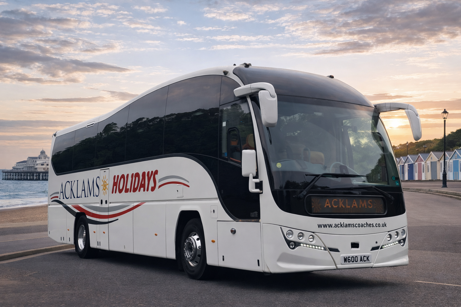 White coach bus with 'ACKLAM'S HOLIDAYS' logo parked on a street near a beach, with colorful beach huts in the background at sunset.