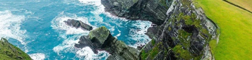 Cliffside with ocean waves crashing against rocks and lush green grass on top.