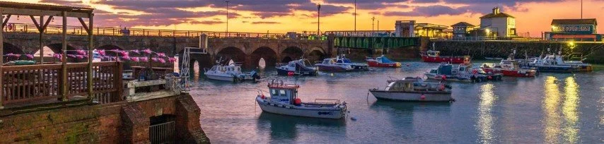 Boats docked in a harbor at sunset with colorful sky, bridge, and buildings in the background.