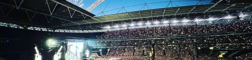 Large stadium filled with spectators, bright lights, and concert stage with a screen, during a concert or event.