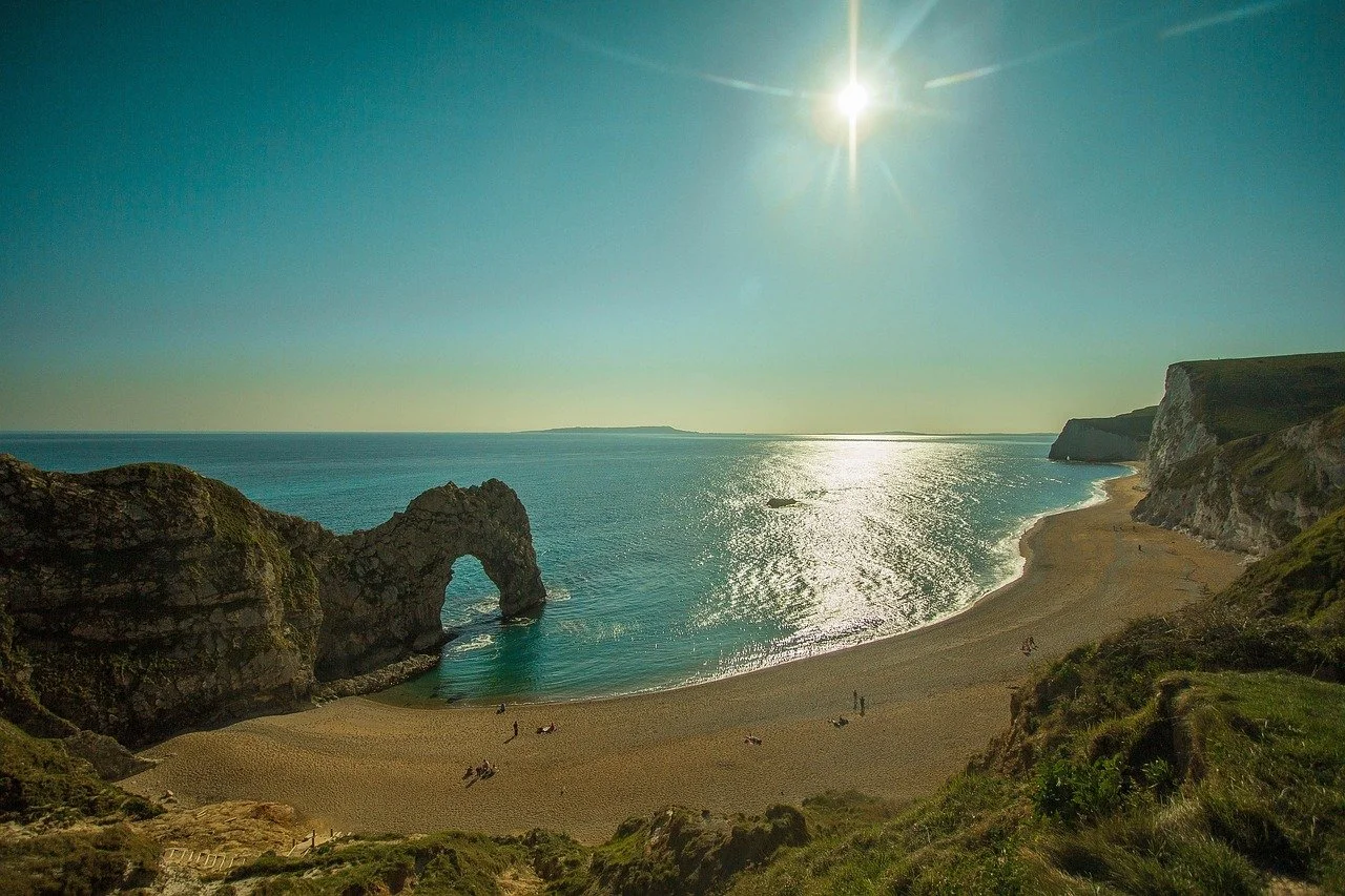 A scenic view of a beach with a large natural rock arch formation in the water, surrounded by cliffs and grassy areas, under a bright sun with clear sky.
