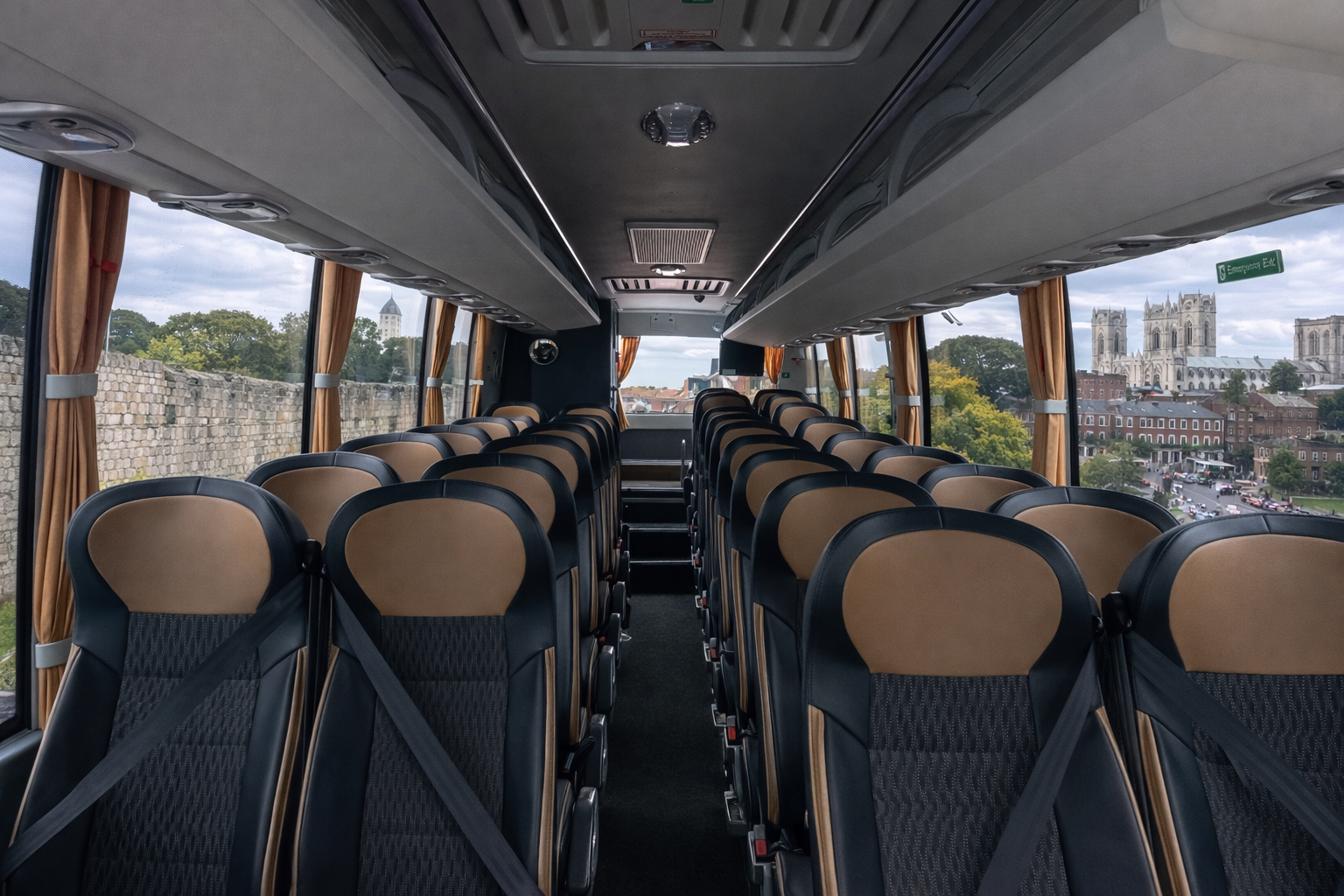Inside a bus with rows of beige and black seats, some seat belts fastened, large windows with orange curtains, and a scenic view of a historic town with cathedral and stone wall outside.