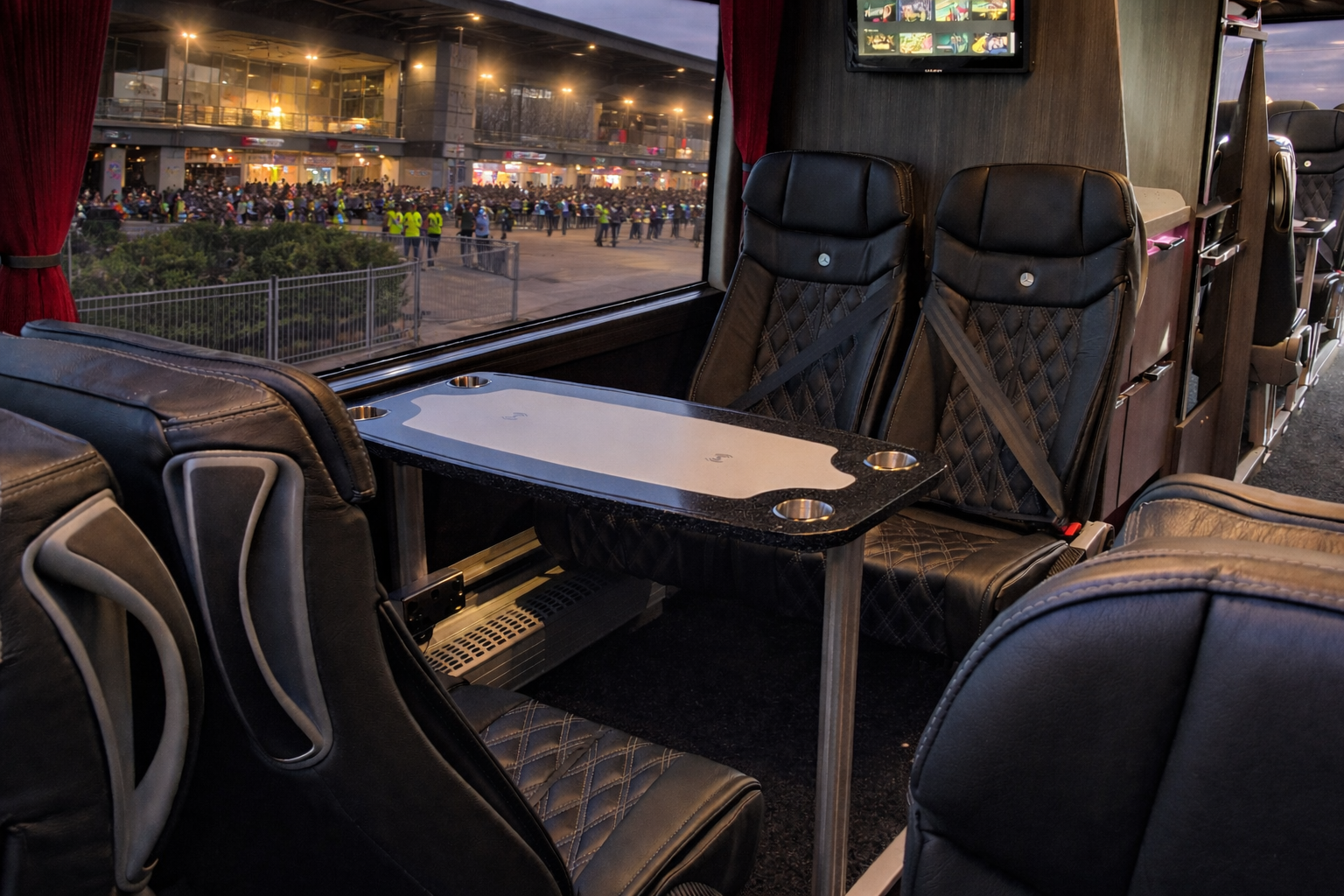 Inside a luxury bus, black leather seats with diamond stitching, a small table with four cup holders, large windows showing a crowd outside, and a television screen mounted on the wall.