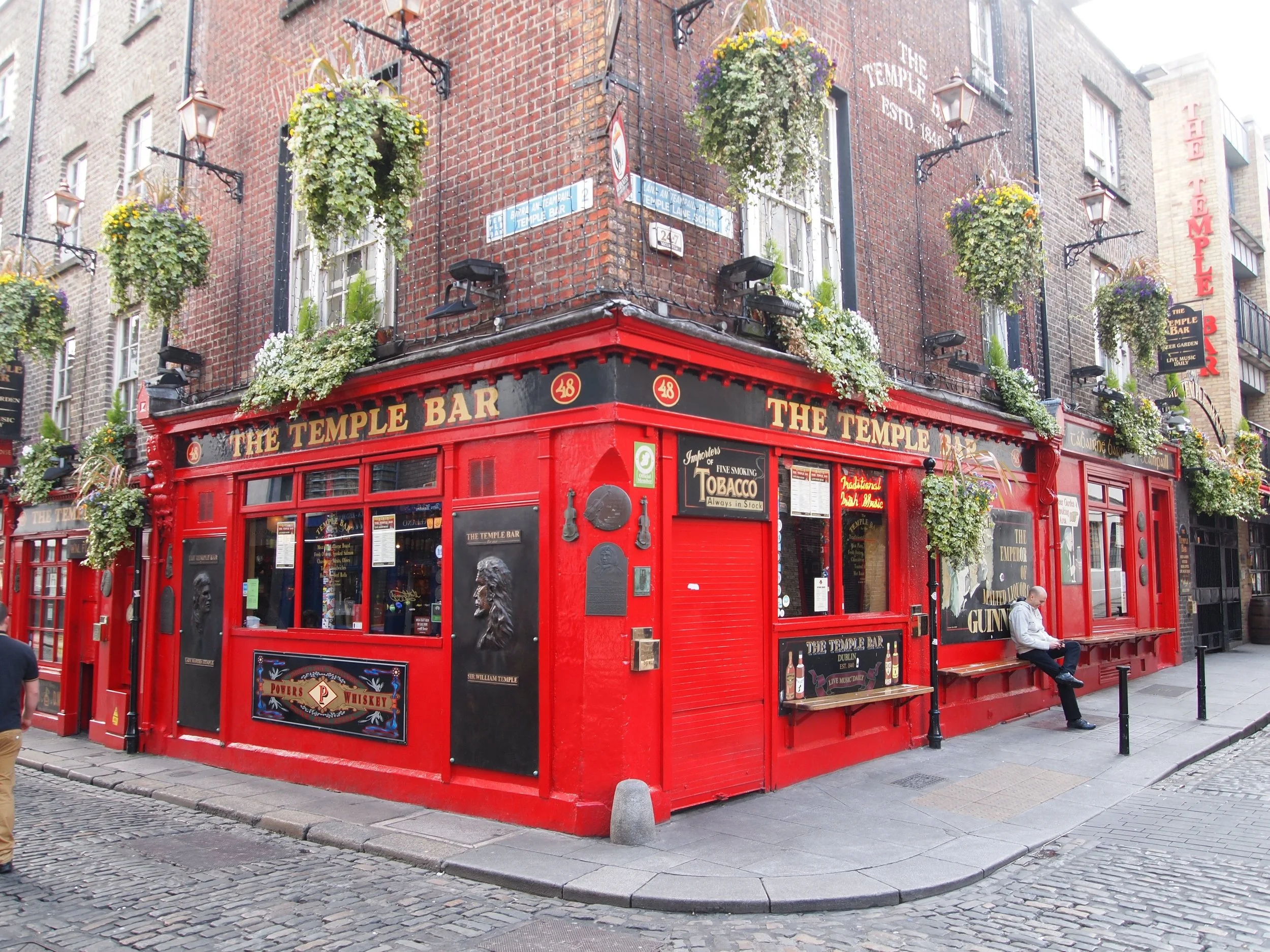 A corner pub called The Temple Bar with red exterior and hanging flower baskets, located on a cobblestone street in Dublin. A man is sitting outside on a bench.