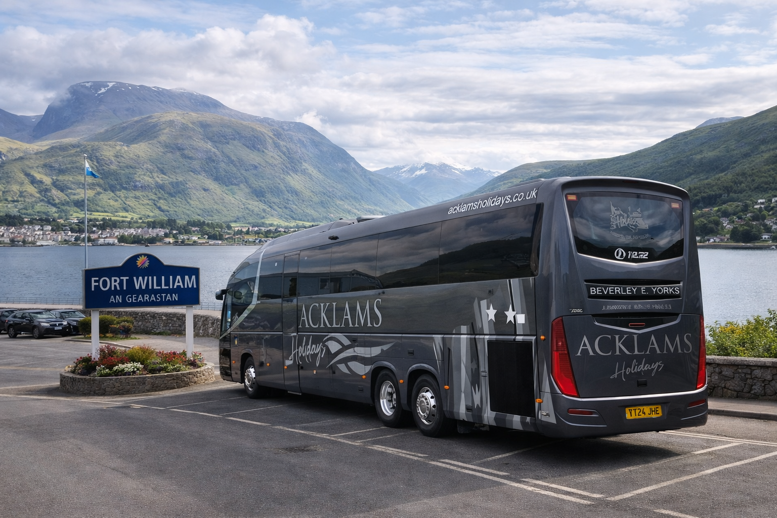 A large gray tour bus with 'ACKLAMS Holidays' written on the side and rear, parked near a lake with mountains in the background. There is a sign that reads 'FORT WILLIAM AN GEARASTAN' and a parking lot with some cars.