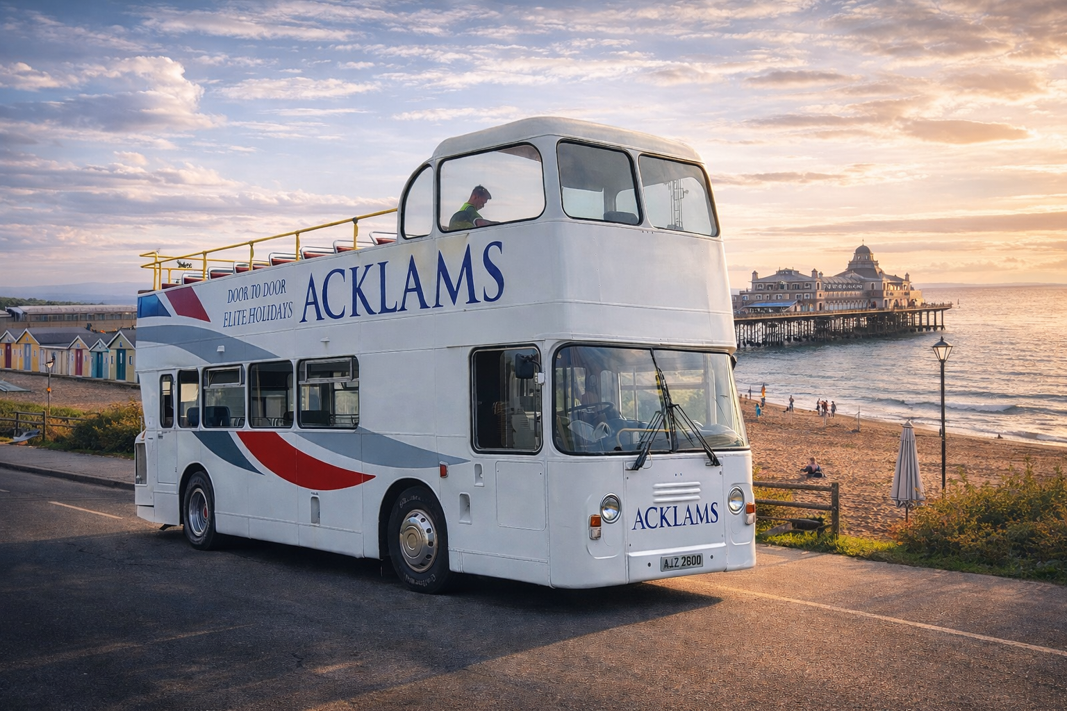 A double-decker bus parked near a beach at sunset with a pier and a large building in the background, along with colorful beach huts and a few people relaxing on the sand.