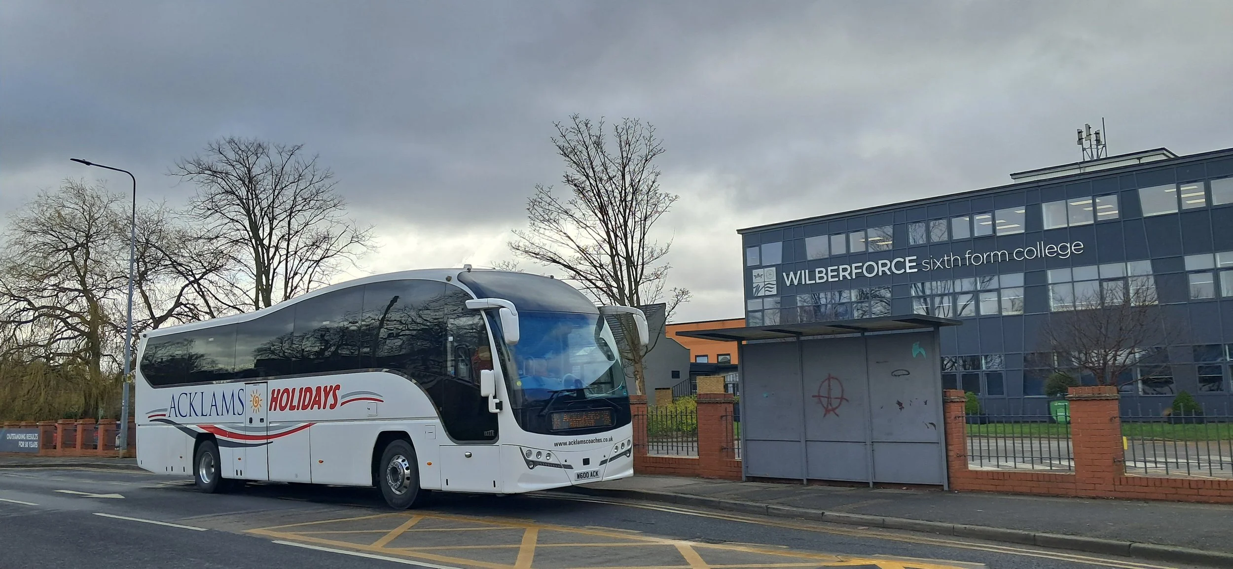 A white coach bus parked on the street near Wilberforce Sixth Form College, with leafless trees and a cloudy sky in the background.