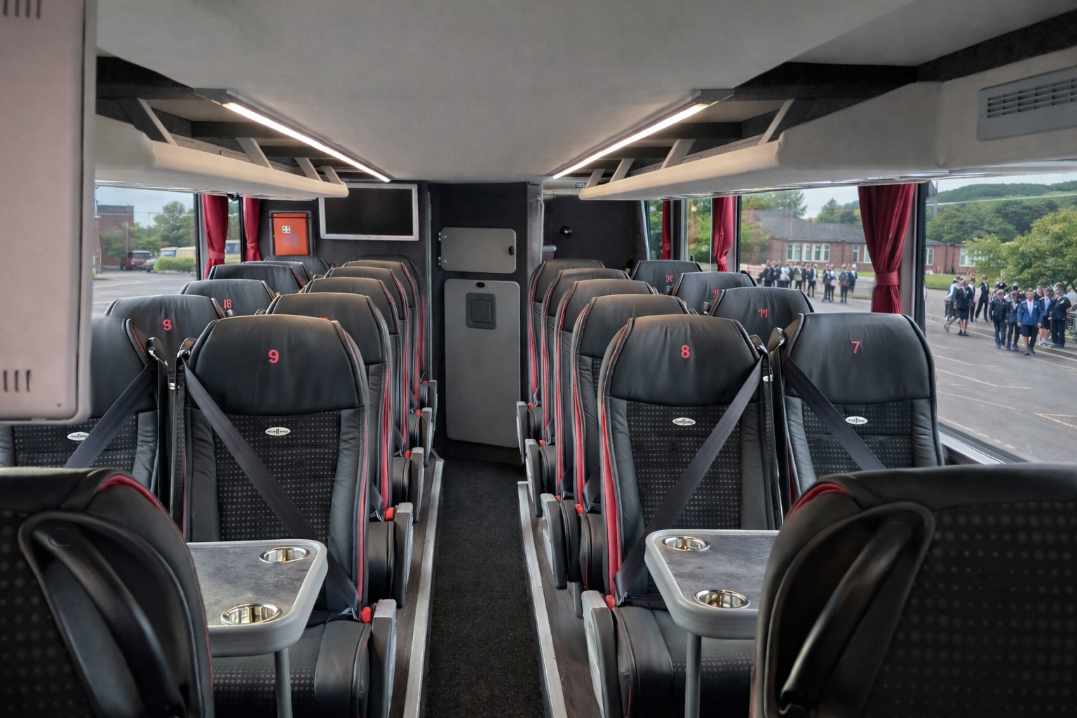 Inside a bus with black leather seats, red numbers, and small foldable tables, looking towards the front with windows showing students in uniforms outside.